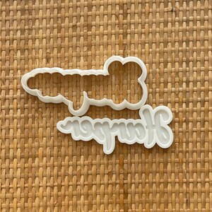 May include: White plastic cookie cutter in the shape of the word "journey" with a second, smaller cutter in the shape of the word "journey" on top of it.
