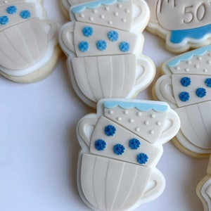 Puede incluir: Galletas decoradas con forma de tazas de té apiladas. Las galletas son blancas con detalles azules, incluyendo decoraciones en forma de flor y bordes festoneados. Las tazas de té están dispuestas en una pila, con un borde azul claro y glaseado blanco.