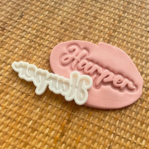 May include: White plastic cookie cutter with the word "Harper" in a cursive font. The cutter is on top of a pink fondant with the same word imprinted in the fondant.