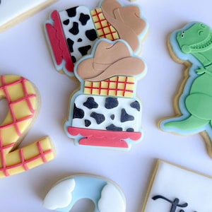 Może przedstawiać: Asortyment zdobionych ciasteczek maślanych w tematyce Toy Story. Ciasteczka obejmują kapelusz kowbojski, wzór w krowy łaty i zielonego dinozaura. Inne ciasteczka mają kształt liczby dwa i mają żółto-różową kolorystykę.