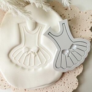 Puede incluir: Un molde de silicona blanco para hacer un adorno de pastel de bailarina. El molde tiene forma de vestido con un lazo y un tutú.