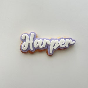 May include: A purple and white cookie with the name "Harper" written in white icing.
