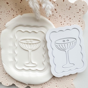 Pode incluir: Um carimbo branco e a sua impressão em argila, ambos com um design de copo de cocktail e uma borda ondulada. O fundo apresenta uma superfície rosa pálido texturizada e elementos decorativos brancos.