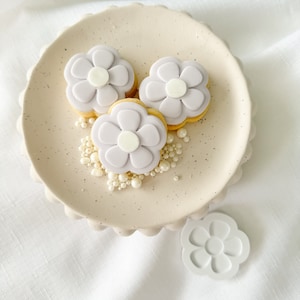 May include: Three purple flower-shaped cookies with white centers on a beige plate with white pearl sprinkles.