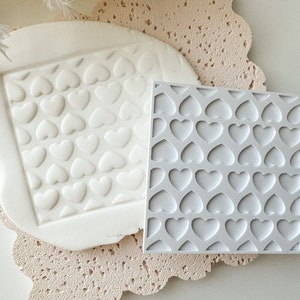 May include: Two white silicone molds for baking, one with a raised heart pattern and the other with a flat heart pattern. Both molds are on a light brown textured surface.
