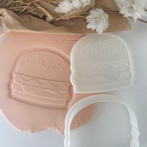 Puede incluir: Cortadores de galletas de plástico blanco con forma de hamburguesa. Los cortadores están sobre una superficie rosa claro.