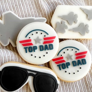 Puede incluir: Una colección de galletas de azúcar decoradas con temática de aviación. Las galletas incluyen gafas de sol de aviador, aviones de combate y galletas redondas con el texto "TOP DAD" y detalles de estrellas en rojo, blanco y azul.