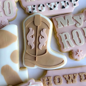 以下が含まれることがあります： ウエスタンをテーマにした、デコレーションされた砂糖クッキーのコレクション。クッキーはカウボーイブーツの形をしており、「Howdy」と「My Rodeo」の文字が白とタン色のアイシングで書かれています。クッキーはピンク、白、タン色のアイシングで飾られています。