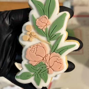 May include: A floral cookie with a white base, decorated with peach-coloured peony flowers and green leaves. The cookie is outlined in yellow and held by a gloved hand. The design includes small, round, yellow accents.