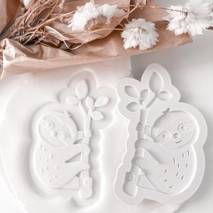 May include: Two white plastic cookie cutters shaped like sloths hanging from a branch. The sloths have smiling faces and are detailed with fur.