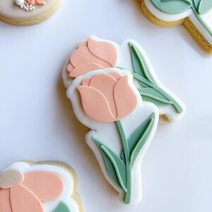 Puede incluir: Tres galletas de az&uacute;car decoradas con glaseado rosa pastel y verde. Las galletas tienen forma de flores con tallo y hojas.