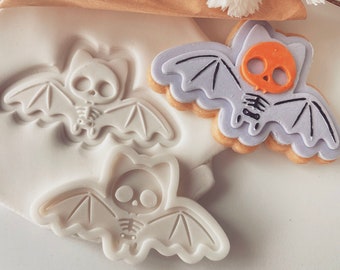 May include: Two white silicone bat cookie cutters and a purple and orange decorated bat cookie. The cookie cutters are in the shape of a bat with a skeleton inside. The decorated cookie is also in the shape of a bat with a skeleton inside and has orange eyes.
