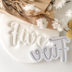 May include: White plastic cookie cutter in the shape of the word "five" with a raised design. The cutter is on top of a white surface with a white plastic impression of the word "five" underneath.