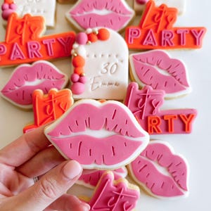 May include: A selection of pink and orange decorated sugar cookies with various designs, including lips, the words "Let's Party", and "Happy 30th Emma".