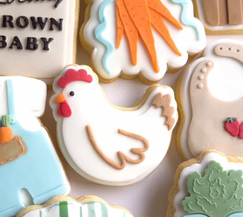Puede incluir: Surtido de galletas de az&uacute;car decoradas. Una galleta de pollo blanco con cresta y pico rojos est&aacute; rodeada de otras galletas, incluyendo un babero con fresas, zanahorias y un traje de beb&eacute;. Las galletas est&aacute;n decoradas con glaseado blanco, naranja, azul y marr&oacute;n.