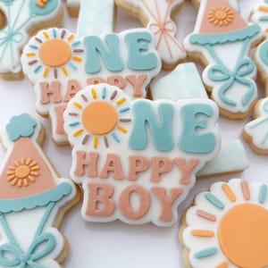 Può includere: Biscotti di zucchero decorati a tema compleanno. I biscotti presentano le parole "ONE HAPPY BOY" in glassa blu e pesca, insieme a disegni di sole e cappello da festa. I biscotti sono decorati con glassa bianca e zuccherini colorati.
