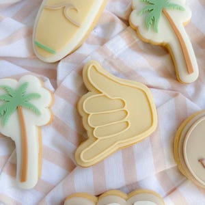 Puede incluir: Surtido de galletas de azúcar decoradas con temática playera. Incluye una tabla de surf, palmeras, un gesto con la mano y galletas con la palabra "RAD". Las galletas son de color amarillo claro con detalles blancos y verdes, dispuestas sobre tela a rayas.