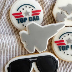 Puede incluir: Surtido de galletas de azúcar decoradas con temática Top Gun. Incluye galletas redondas con el texto "TOP DAD", un avión gris y gafas de sol de aviador. Las galletas presentan detalles en rojo, azul y gris.