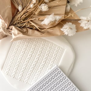 May include: Two white silicone cake decorating stamps with raised designs. One stamp has a woven pattern and the other has a repeating leaf pattern.