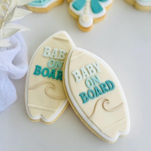 Puede incluir: Dos galletas con forma de tabla de surf con el texto "BABY ON BOARD" en azul claro y turquesa. Las galletas están decoradas con glaseado blanco y un diseño de olas. Otras galletas con temática de bebé están en el fondo.