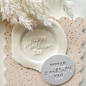 以下が含まれることがあります： 「Happy Mother's Day」という文字が書かれた白いシリコンモールドが2つ。モールドは薄いピンクのテクスチャのある表面の上に置かれています。