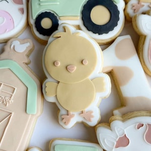 May include: A selection of decorated sugar biscuits with a farm animal theme. The biscuits include a yellow chick, a green tractor, a cow, a barn, and the number one. The biscuits are decorated with pastel colours and white icing.