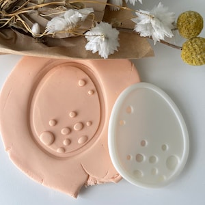 May include: A light pink clay egg shape with a pattern of circles pressed into it, next to a white plastic egg-shaped cutter with matching holes. Dried flowers and foliage are in the background.