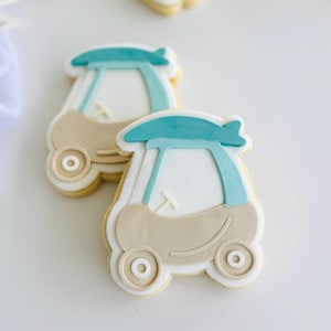 May include: Two car-shaped sugar cookies. The cookies are decorated with light brown, white, and teal icing. The cookies have a car-like design with wheels, a windshield, and a roof. The cookies are on a white surface.