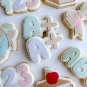 May include: A set of nine decorated sugar cookies featuring a back to school theme. The cookies are decorated with pastel colors and feature numbers, letters, a bow, a stack of books, and an apple.