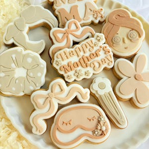 Op de afbeelding: Een set van negen gedecoreerde suikerkoekjes in een lichtbruin en wit kleurenschema. De koekjes hebben verschillende ontwerpen, waaronder een bloem, een strik, een slaapmasker, een haarspeld, een vrouwen gezicht en de woorden "Happy Mother's Day" en "Mama".