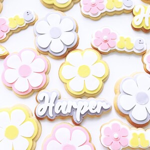 May include: A set of decorated sugar cookies featuring a daisy design in various colours, including pink, yellow, and white. The cookies also include the name "Harper" and the word "one" in a playful font.