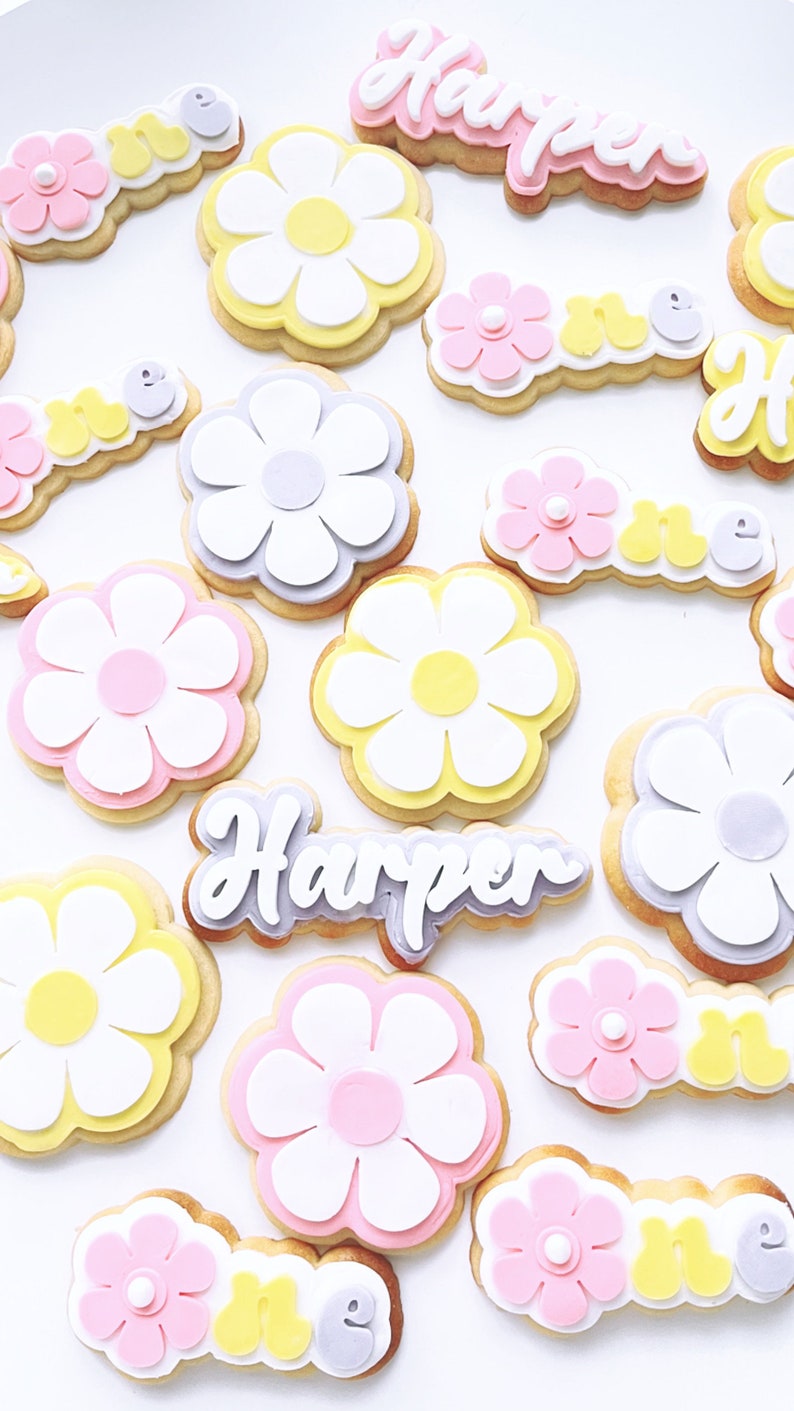 May include: A set of decorated sugar cookies featuring a daisy design in various colours, including pink, yellow, and white. The cookies also include the name "Harper" and the word "one" in a playful font.