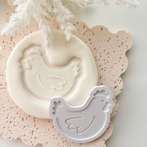 Puede incluir: Un molde de silicona blanco y un cortador de galletas gris, ambos con forma de gallina, se exhiben sobre una superficie texturizada rosa claro con bordes festoneados. Elementos decorativos blancos y plumosos est&aacute;n en el fondo. Los art&iacute;culos son probablemente para hornear.