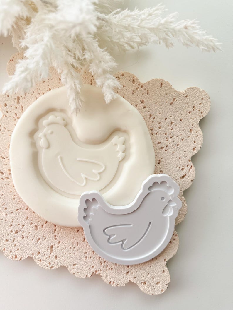 Puede incluir: Un molde de silicona blanco y un cortador de galletas gris, ambos con forma de gallina, se exhiben sobre una superficie texturizada rosa claro con bordes festoneados. Elementos decorativos blancos y plumosos est&aacute;n en el fondo. Los art&iacute;culos son probablemente para hornear.