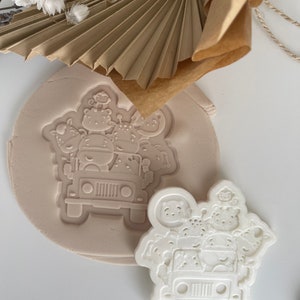 May include: A white plastic cookie cutter shaped like a jeep with animals inside. The cutter is on a light brown surface.