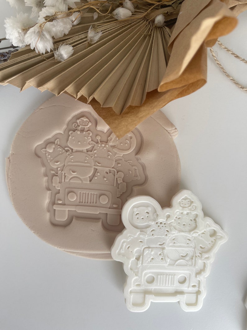 May include: A white plastic cookie cutter shaped like a jeep with animals inside. The cutter is on a light brown surface.