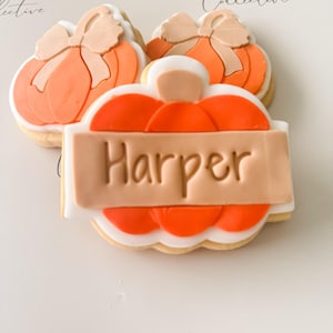 May include: Three pumpkin-shaped cookies with orange and tan frosting. One cookie has a rectangular tan banner with the name "Harper" in brown lettering. The cookies are decorated with white and orange details, and a bow.
