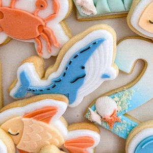Könnte beinhalten: Eine Sammlung von dekorierten Zuckerkeksen mit einem blauen Wal, einem orangefarbenen Krebs, einem gelben Fisch und einer Muschel. Die Kekse sind mit weißem Zuckerguss und blauen, orangefarbenen und gelben Details verziert.