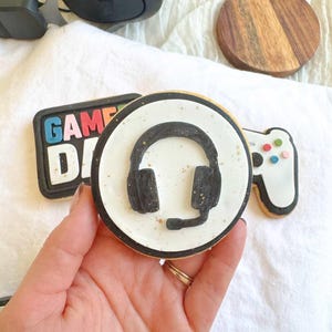 Peut inclure: Ensemble de biscuits au sucre décorés sur le thème des jeux vidéo. L'un des biscuits présente un motif de casque audio, un autre affiche "GAME DAY" en lettres colorées, et un troisième a la forme d'une manette de jeu. Un casque noir et un dessous de verre en bois sont également visibles.