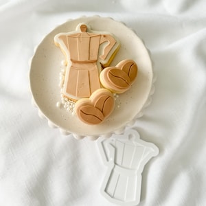 Op de afbeelding: Een wit keramiek bord met een bruine suikerkoek in de vorm van een koffiekan en twee bruine suikerkoekjes in de vorm van koffiebonen. Het bord staat op een wit oppervlak.