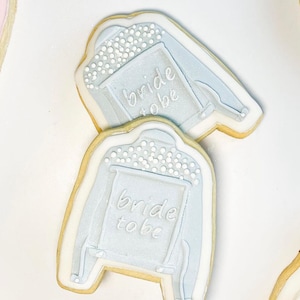May include: Two light blue cookies shaped like jackets, decorated with white icing and small white sprinkles. The cookies have the words "bride to be" written on them in white icing. The cookies are on a white surface.