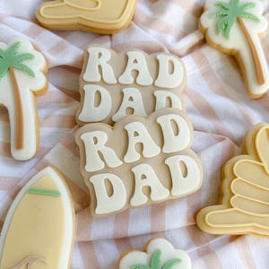 Puede incluir: Galletas decoradas con temática playera. Las galletas incluyen una tabla de surf, palmeras, un gesto con la mano y galletas que deletrean "RAD DAD" en glaseado blanco sobre un fondo beige. Las galletas están dispuestas sobre una tela a rayas.