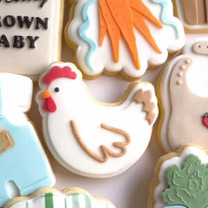 Puede incluir: Surtido de galletas de az&uacute;car decoradas. Una galleta de pollo blanco con cresta y pico rojos est&aacute; rodeada de otras galletas, incluyendo un babero con fresas, zanahorias y un traje de beb&eacute;. Las galletas est&aacute;n decoradas con glaseado blanco, naranja, azul y marr&oacute;n.