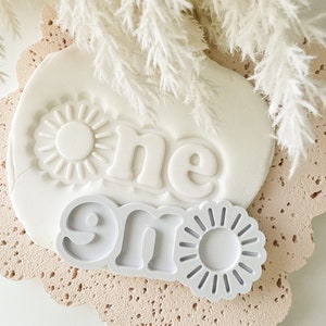 May include: White clay mould with the word "one" embossed in a stylised font. The mould is surrounded by white pampas grass.