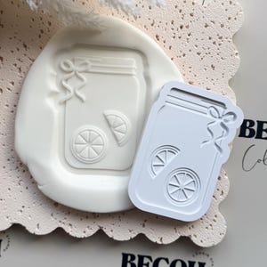 Könnte beinhalten: Weißer Keksstempel und Abdruck eines Einmachglases mit Zitronenscheiben und einer Schleife. Der Stempel liegt neben dem Abdruck auf einer strukturierten, hellfarbigen Oberfläche. Der Text "BECOH Collective" ist schwarz gedruckt.