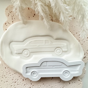 Puede incluir: Dos moldes de silicona blancos de coches clásicos. Los moldes están sobre una superficie texturizada rosa claro.