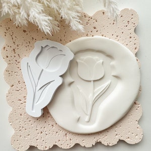 Puede incluir: Molde de silicona blanco con un dise&ntilde;o de tulip&aacute;n. El molde est&aacute; sobre una superficie texturizada rosa claro. Un sello de tulip&aacute;n de pl&aacute;stico blanco est&aacute; al lado del molde.