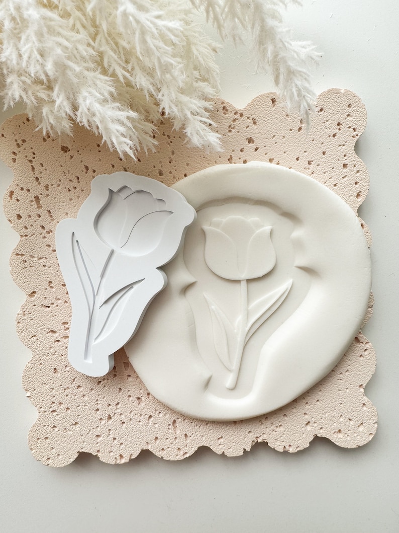 Puede incluir: Molde de silicona blanco con un dise&ntilde;o de tulip&aacute;n. El molde est&aacute; sobre una superficie texturizada rosa claro. Un sello de tulip&aacute;n de pl&aacute;stico blanco est&aacute; al lado del molde.