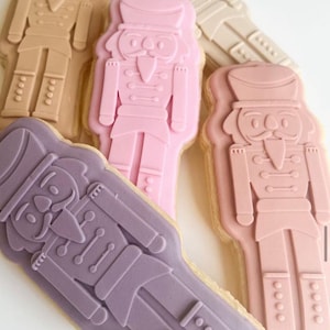 May include: Four decorated sugar cookies shaped like nutcracker soldiers. The cookies are decorated with pink, purple, and brown icing.