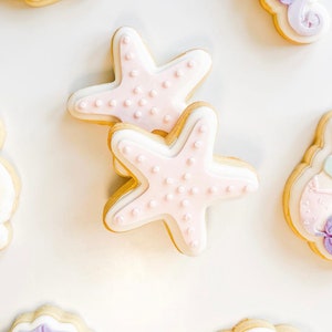 Puede incluir: Dos galletas de azúcar rosa y blancas decoradas con un diseño de estrella de mar. Las galletas tienen un fondo blanco con puntos rosas.